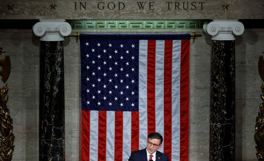 Spreker Mike Johnson staat op een podium voor een grote Amerikaanse vlag en onder een inscriptie "Op God vertrouwen wij"