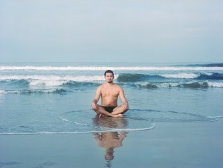 Shirtless person sitting cross-legged in shallow water at a beach.