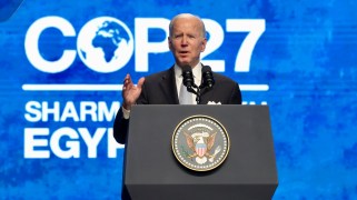 Joe Biden in a suit speaking behind a podium with the text "COP 27" behind him on a blue background