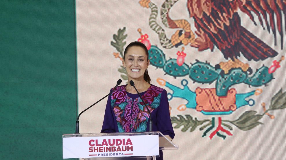 Claudia Sheinbaum, standing at a podium with her name on it, in front of a large Mexican flag