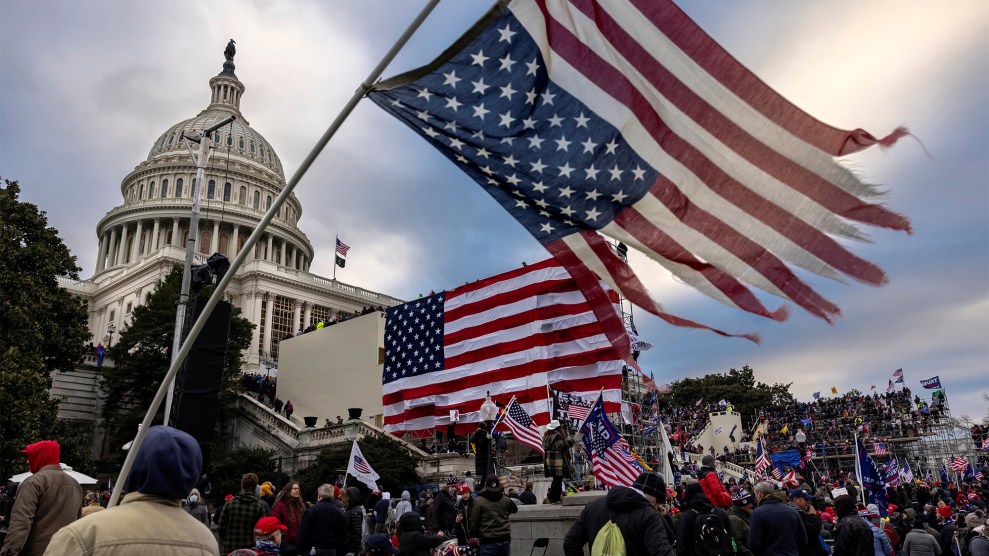 A crowd gathers in front of the US Capitol. A tattered American flag flies in the foreground.