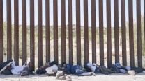 Migrants lying down against a tall fence at the US border