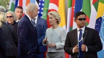 President Joe Biden talks to European Commission President Ursula von der Leyen in front of multi-colored flags of various countries at the G7 summit in Italy.