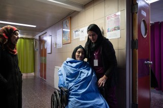 Woman with her eyes closed, sitting in a wheelchair in a hospital. Another woman is standing next to her. A third woman looks on from the left of the frame.