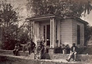 Black and white photo of four men sitting in front of a small building.