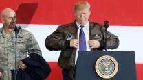 The image shows former President Donald Trump standing at a podium with the presidential seal, wearing a leather bomber jacket. The jacket has a patch that reads "Donald J. Trump Commander in Chief." He appears to be in the process of adjusting or zipping up the jacket. Next to him is a man in a military uniform.