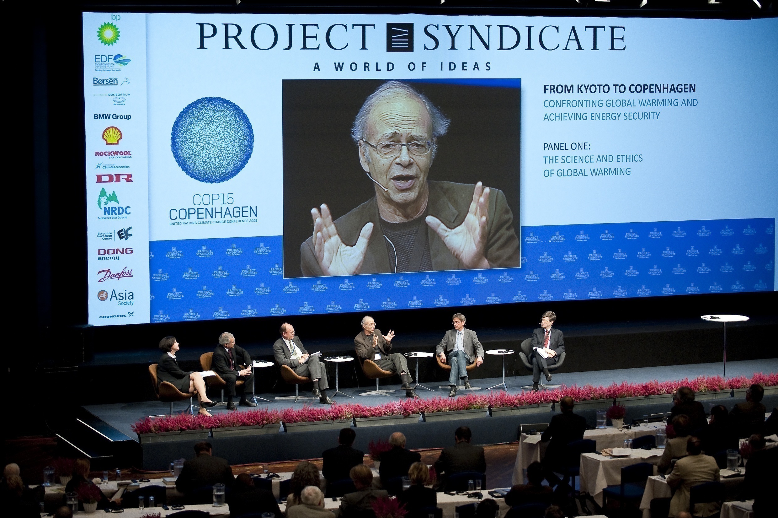 Row of six speakers on a stage with a large screen above them showing Peter Singer speaking.