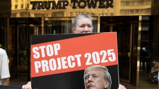 A white person standing in front of the Trump Tower with a poster that says "Stop Project 2025" that has a photo of Donald Trump on it.