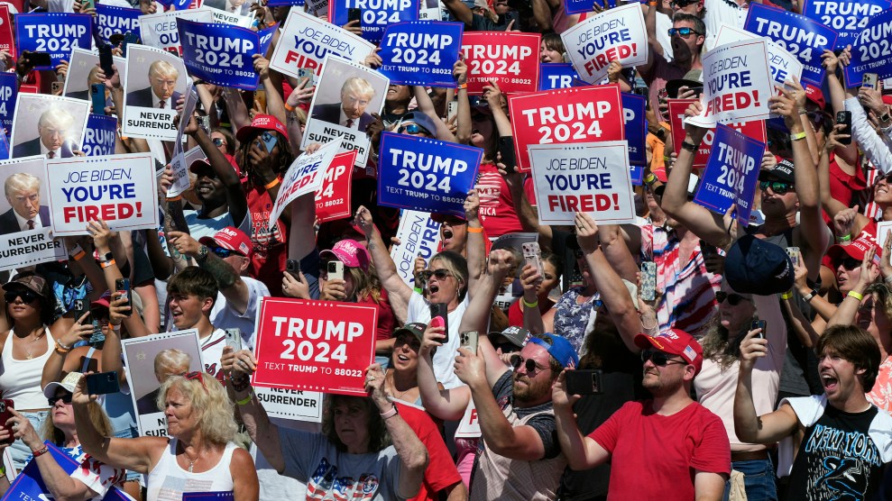 Attendees at a Trump rally, some of whom look sunburned, holding up picture of Trump's mugshot and signs that says "Trump 2024" and "Joe Biden, You're Fired"