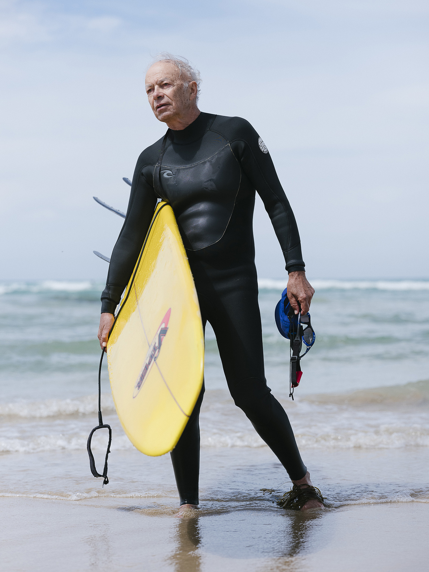 Peter Singer wears a wetsuit while standing on a beach, holding a surfboard.