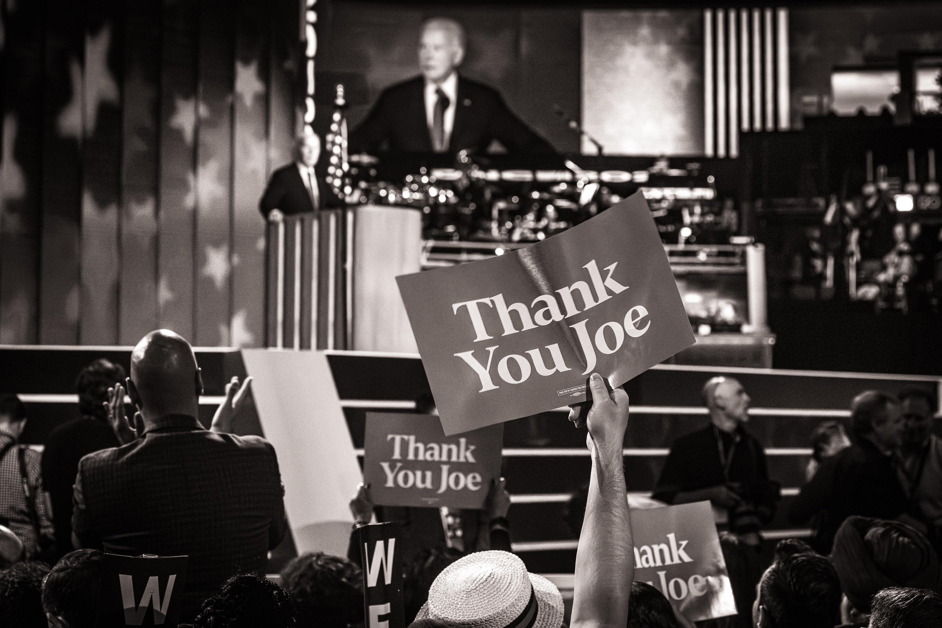 Joe Biden speaking at a podium. Members of the audience hold signs that say "Thank You Joe."