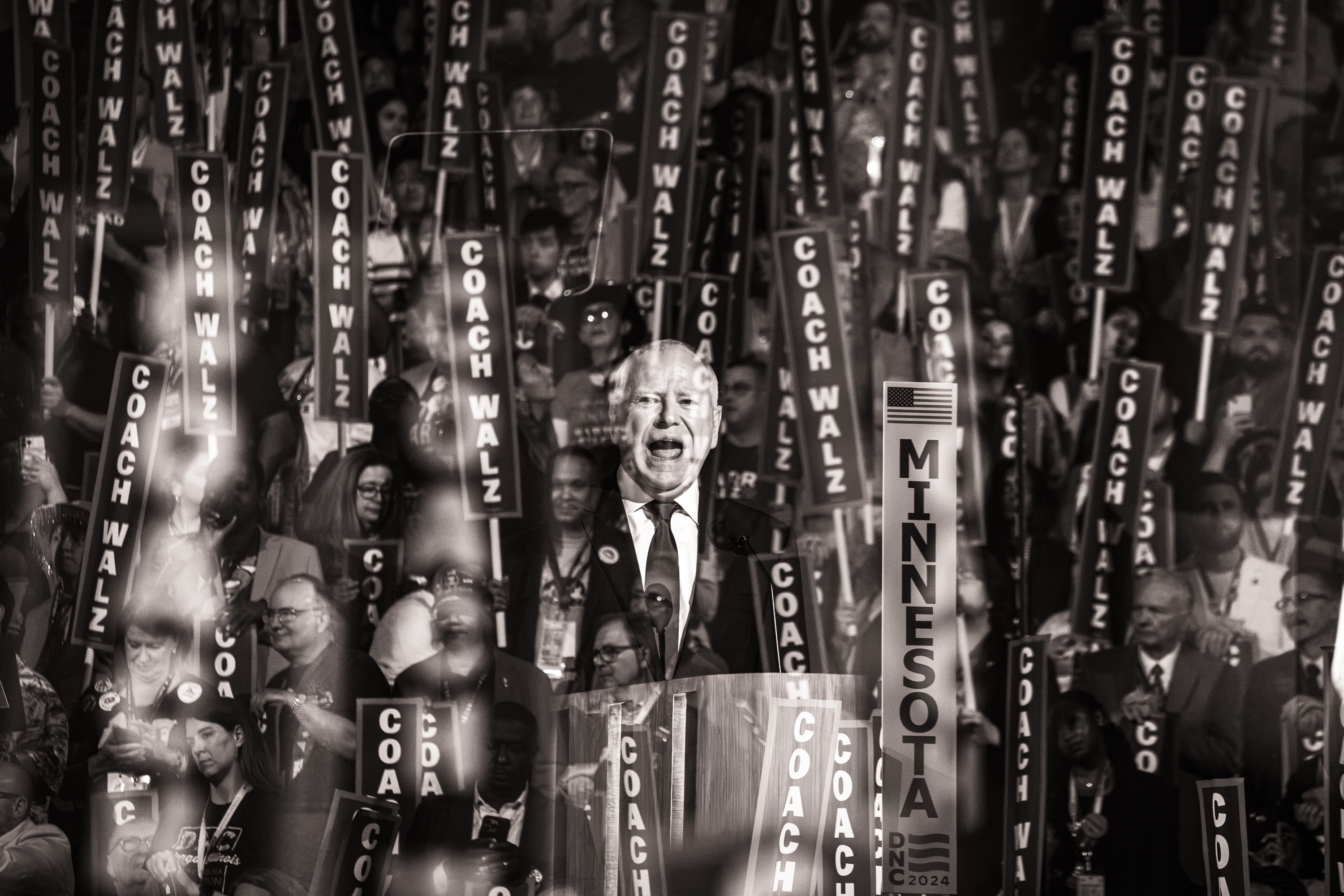 Tim Walz hablando ante una multitud con carteles que dicen "Entrenador Walz".