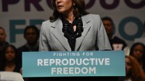 Kamala Harris standing behind a podium during a public event. The podium has a sign that reads "FIGHTING FOR REPRODUCTIVE FREEDOM" in bold, white letters against a teal background. Kamala Harris is dressed in a light gray blazer and is speaking into two microphones.