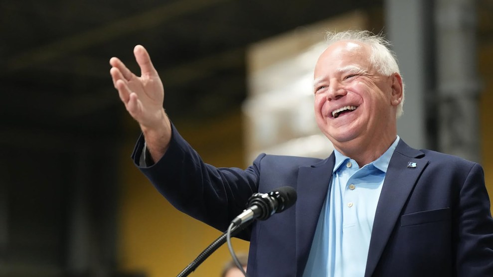 Tim Walz in a suit and at a podium gesturing to the crowd with a large smile on his face.