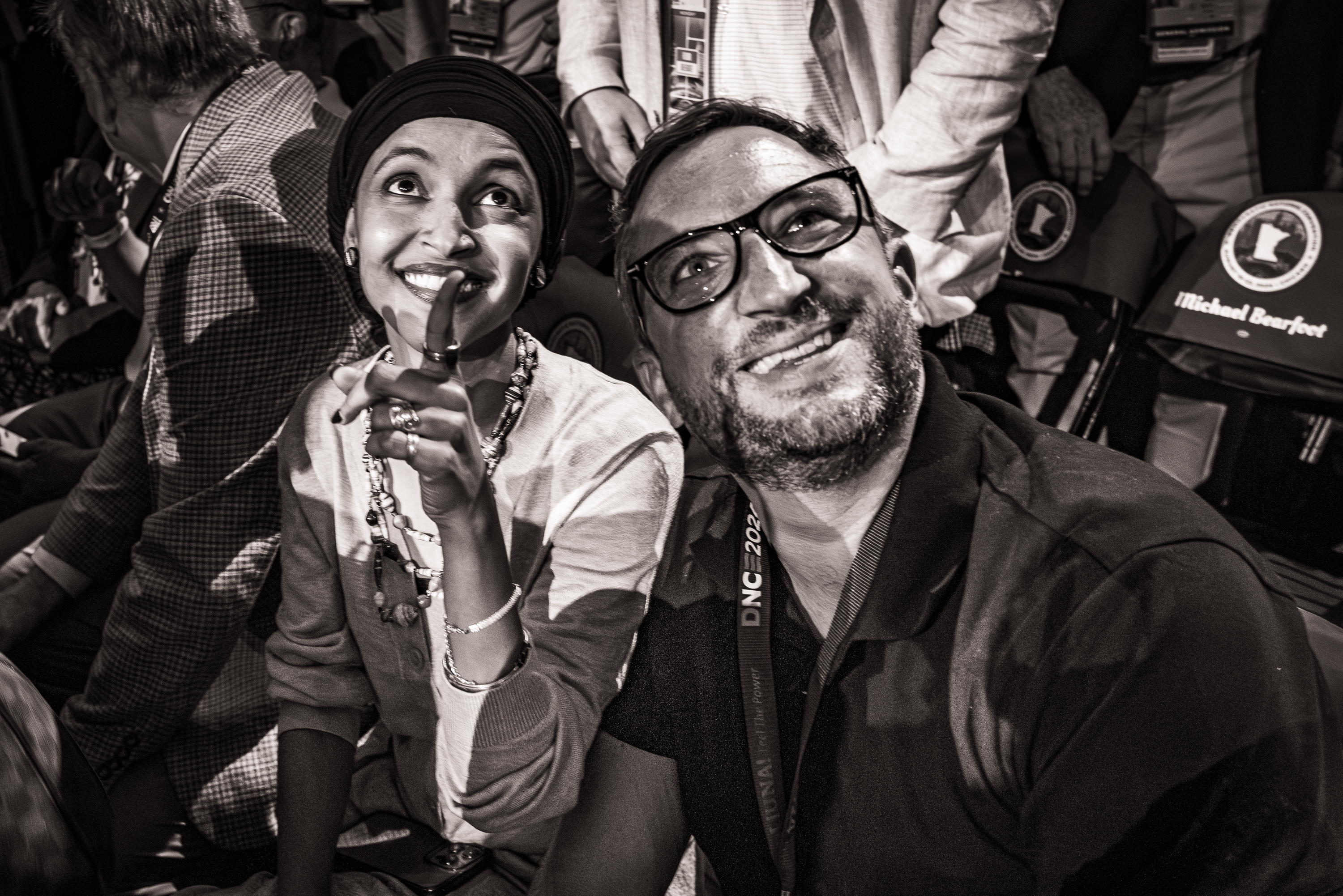 Black and white photo of Representative Ilhan Omar and a man looking up, smiling and pointing.