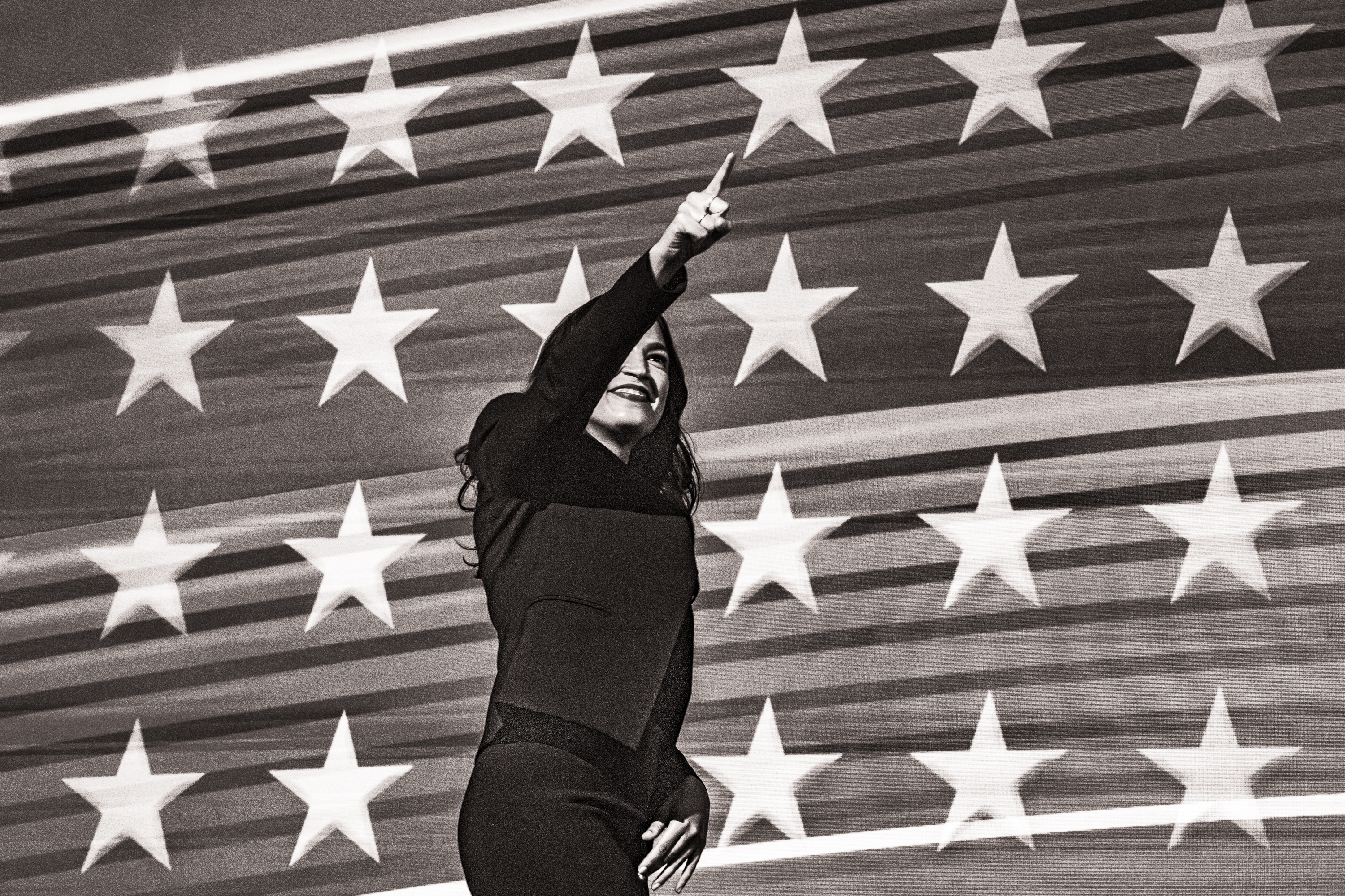 Black and white photo of Representative Alexandria Ocasio-Cortez pointing and smiling as she comes on stage at the DNC.