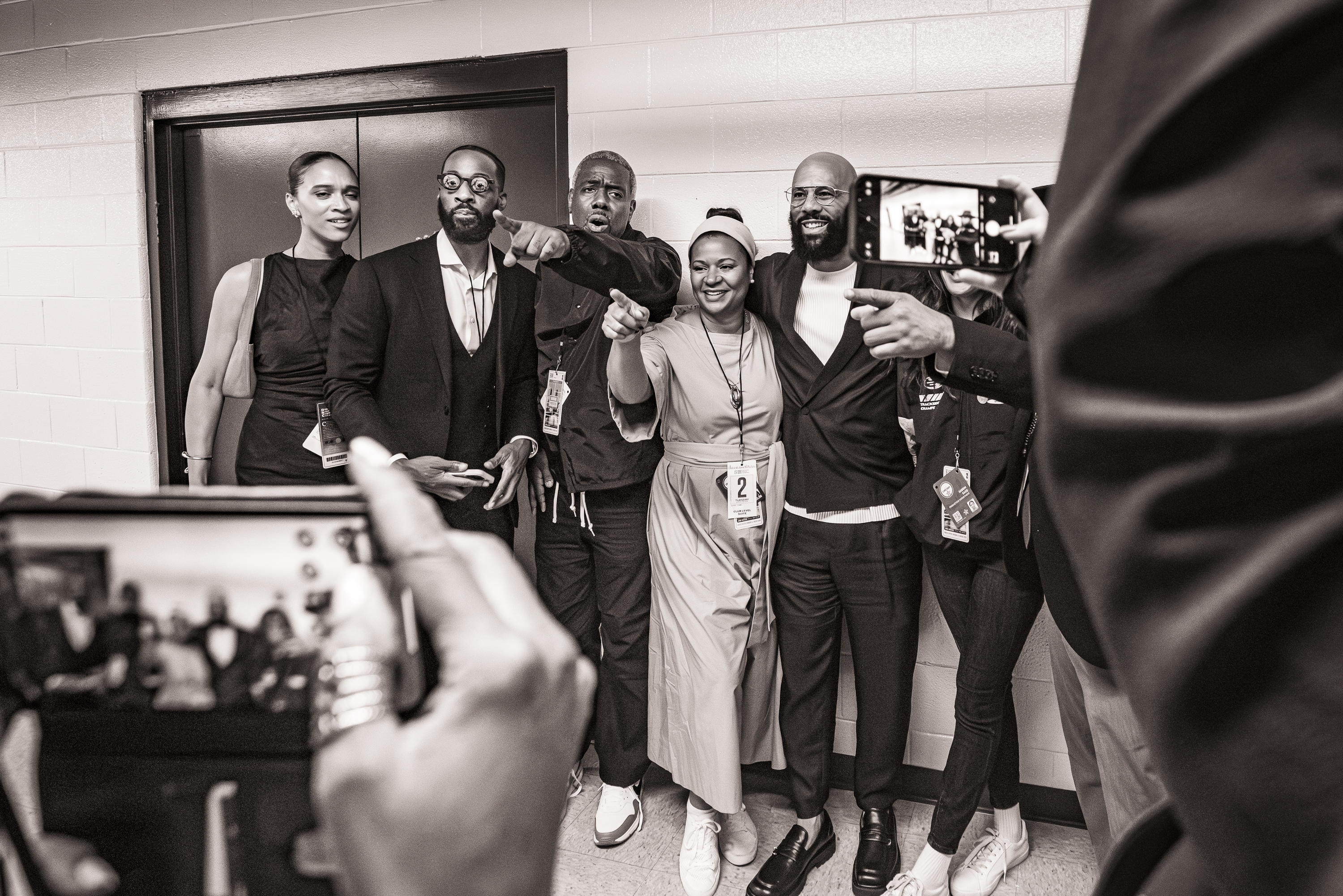 A Black and white photo of a group of people stand together for a photo, with people holding cellphones in the foreground.