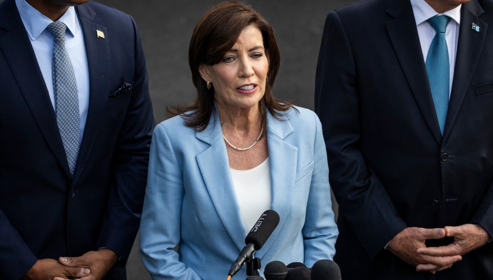 New York Governor Kathy Hochul is a white woman with brown hair wearing a light blue suit. She is speaking into a microphone and behind her are two men in suits, one white and one black.