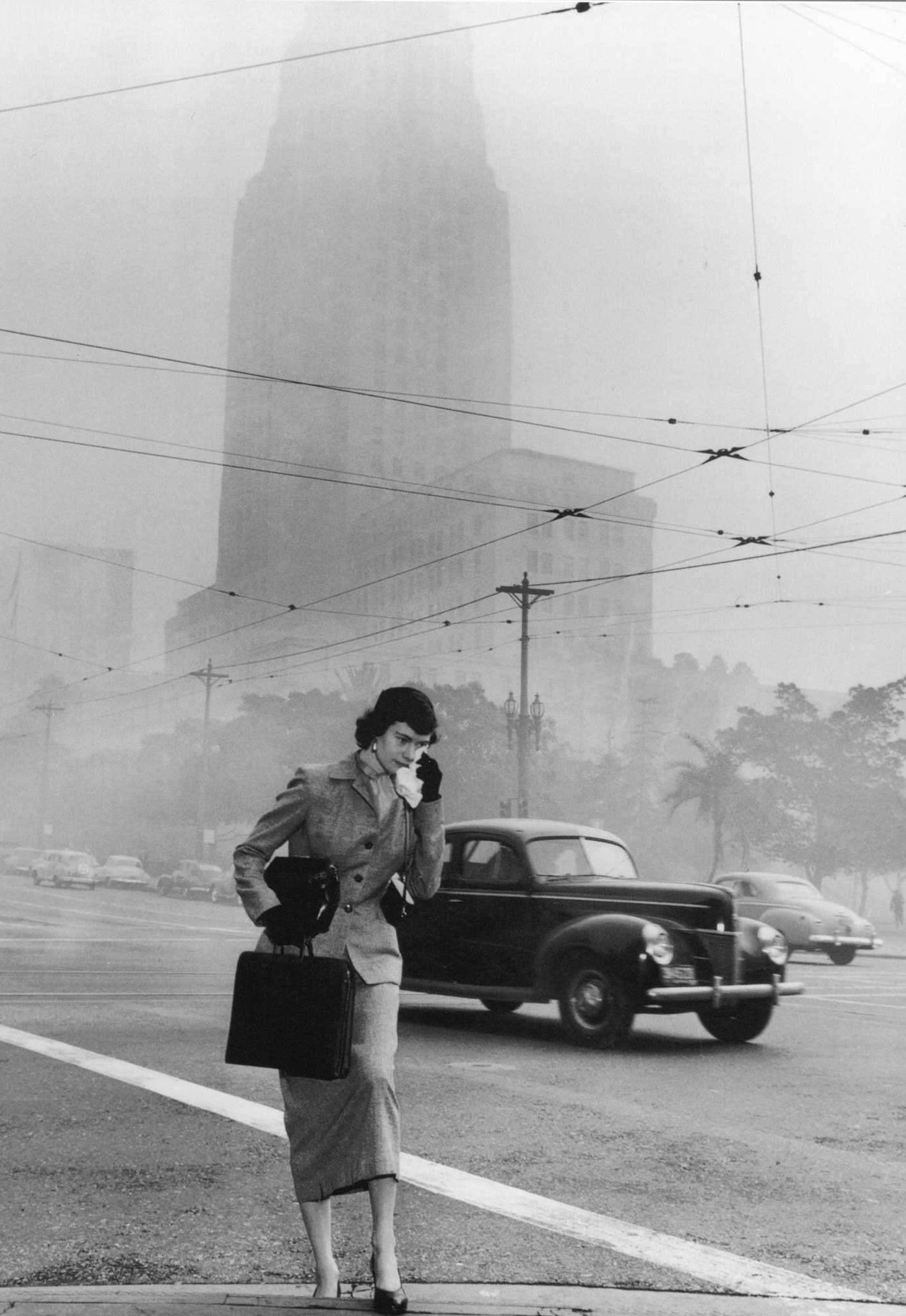 Black and white photo of a woman blotting her eyes with a handkerchief while walking through a smoggy intersection.