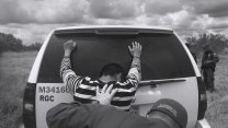 A black-and-white photograph shows a young man with his hands pressed against the back of an SUV, being searched by a U.S. Customs and Border Protection agent. The young man is wearing a striped shirt and appears to be looking down. The agent, wearing a cap and gloves, is seen from behind as he performs the search. Another agent stands nearby, observing the situation. The scene is set in an open, rural area with tall grass and scattered bushes under a cloudy sky.