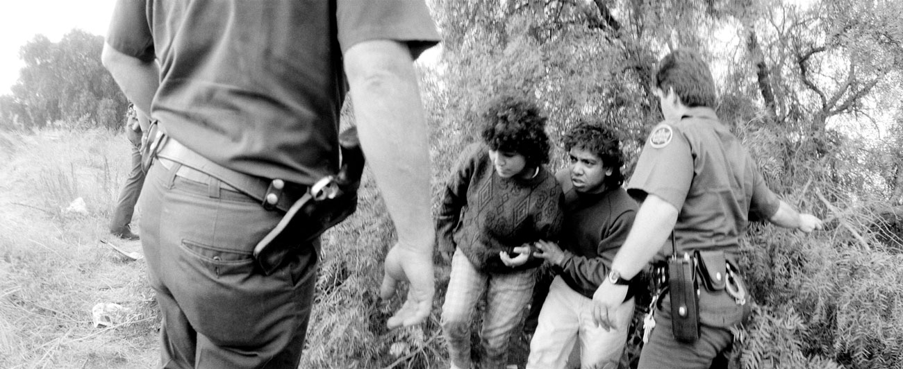 A black-and-white photograph shows two young men being apprehended by border patrol agents. The young men appear distressed, huddling together near some bushes. One agent is seen from behind, approaching the scene with a hand extended, while another agent holds one of the young men by the arm. The agents are in uniform, with visible badges and utility belts. The scene is set in a rural, overgrown area with bushes and tall grass.