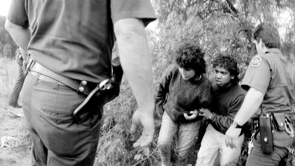 A black-and-white photograph shows two young men being apprehended by border patrol agents. The young men appear distressed, huddling together near some bushes. One agent is seen from behind, approaching the scene with a hand extended, while another agent holds one of the young men by the arm. The agents are in uniform, with visible badges and utility belts. The scene is set in a rural, overgrown area with bushes and tall grass.