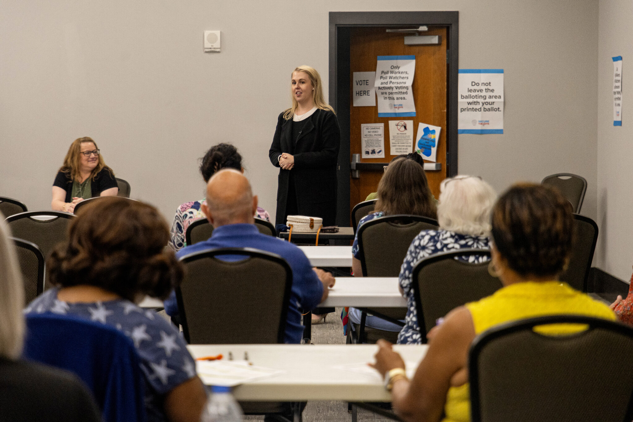 A blonde woman wearing black faces three rows of people seated at folding tables.