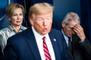 Donald Trump out of focus in the foreground with Dr. Deborah Birx behind him to the left and Dr. Anthony Fauci to his rigth, with his head in his hand.