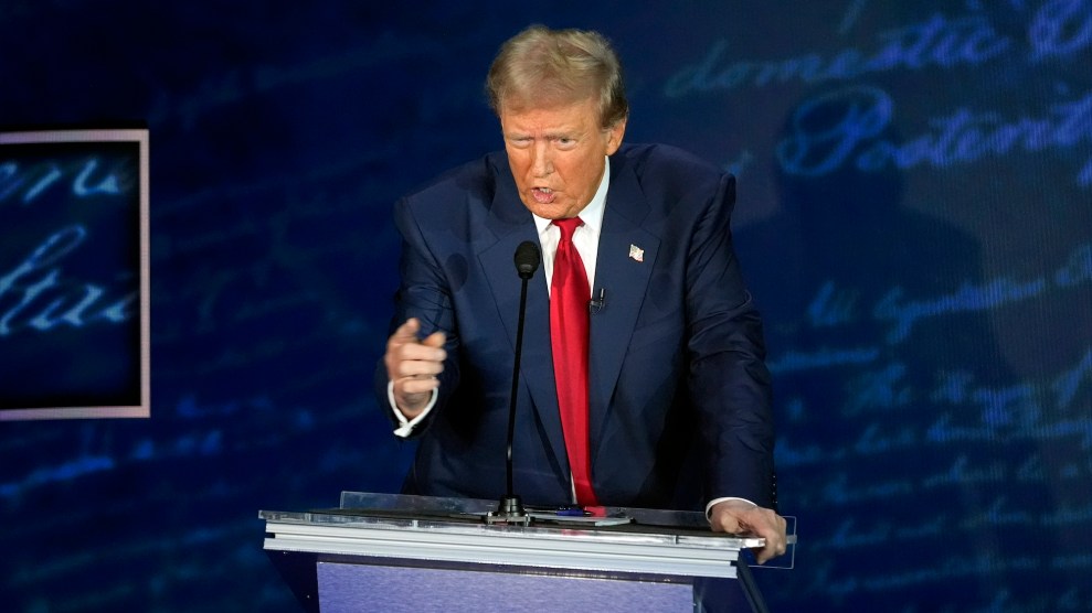 Donald Trump in a blue suit and a red tie pointing ahead while speaking behind a podium.