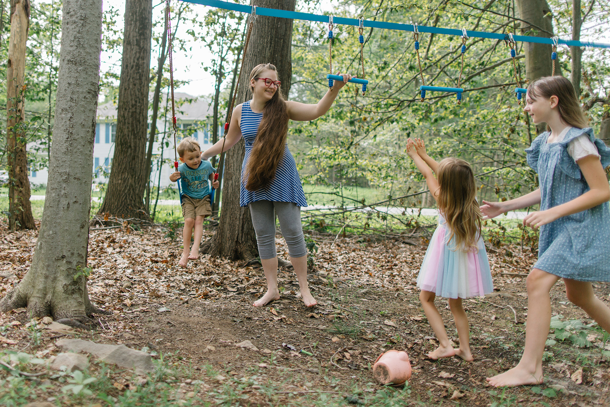 Grace Smith, a White woman wearing glasses, a blue and white striped tank top and gray leggings, pushes one child on a swing while talking to her other children.