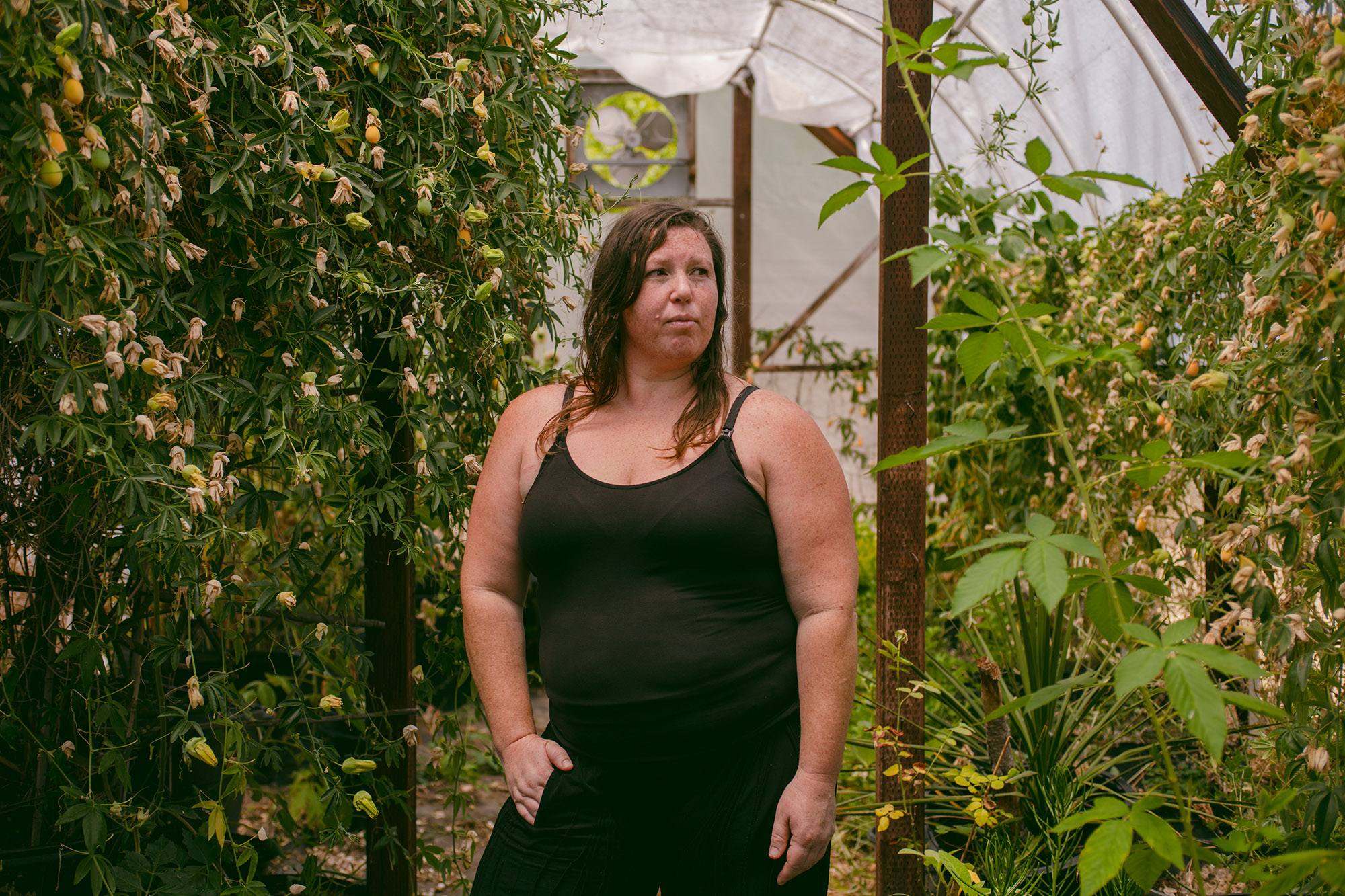 Susan Horton, a white woman wearing a black tank top and black pants, poses for a portrait inside a greenhouse.