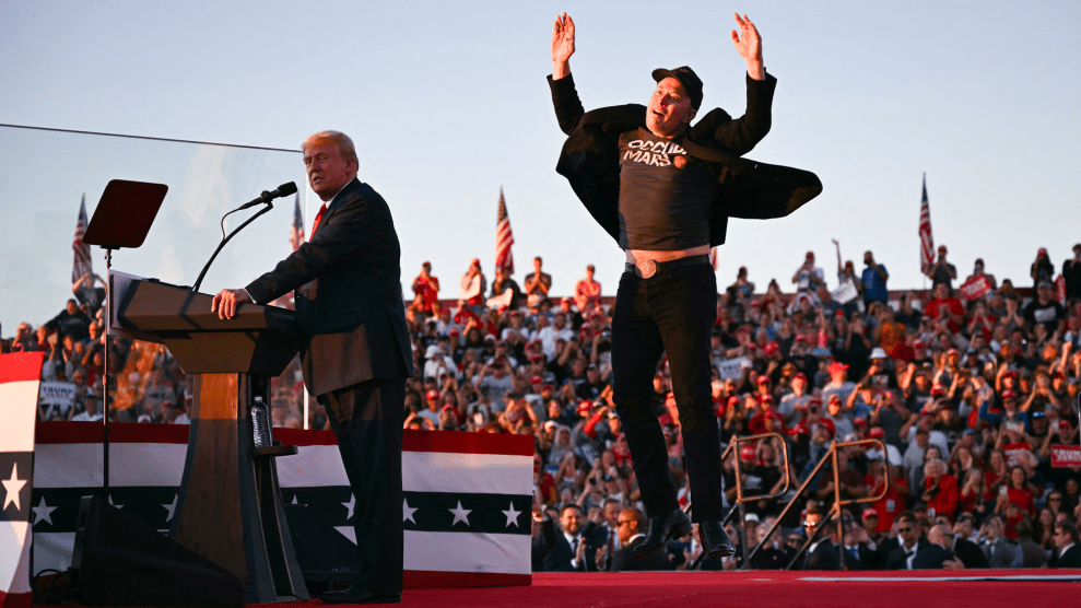 Elon Musk, dressed all in black, on stage behind Donald Trump. As Musk comes down from his jump, his arms are raised in the air, his jacket flapping in the wind.