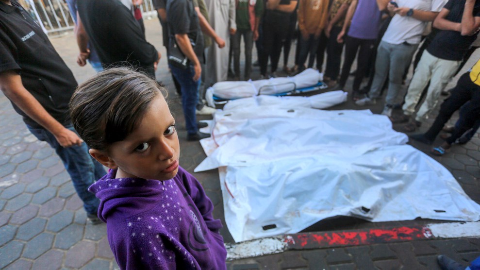 A child dressed in a purple hoodie stars into the camera. Behind the child are people standing around multiple bodies lying on the ground covered in white tarps.