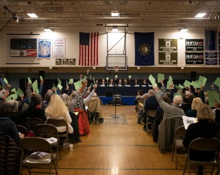 School gymnasium filled with people sitting in folding chairs, some with their arms raised, holding pieces of paper.