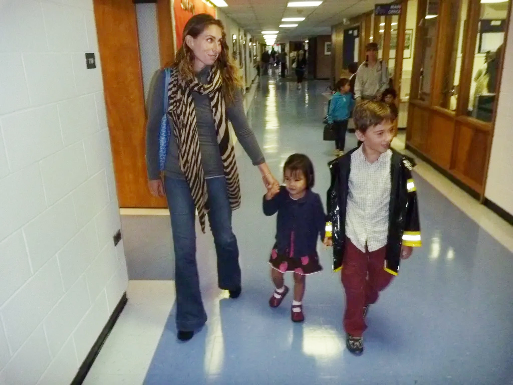 A woman wearing a long animal-print scarf walks down a school hallway, holding the hand of her young daughter, who wears a navy dress with pink apples on it. The girl also holds hands with her older brother.