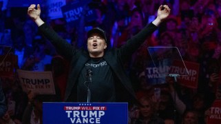 Elon Musk has his hands in the air in front of a microphone and a Trump/Vance sign