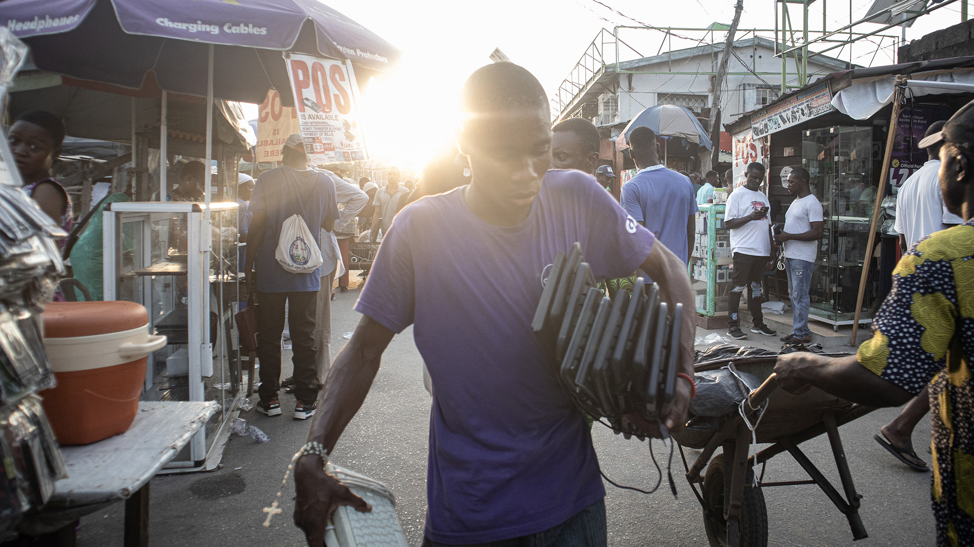 Joven cargando varios dispositivos electrónicos en el mercado de Ikeja Computer Village en Lagos, Nigeria. El mercado es un centro caótico de comercio y reciclaje de residuos electrónicos, donde se reutilizan y desmontan dispositivos obsoletos.