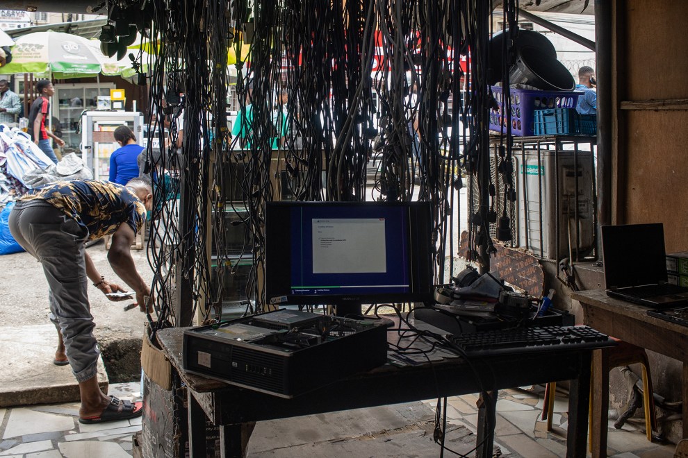 Taller en Ikeja Computer Village con un ordenador en funcionamiento y cables colgando del techo. En el fondo, un hombre recolecta residuos electrónicos mientras los negocios operan ajetreadamente.