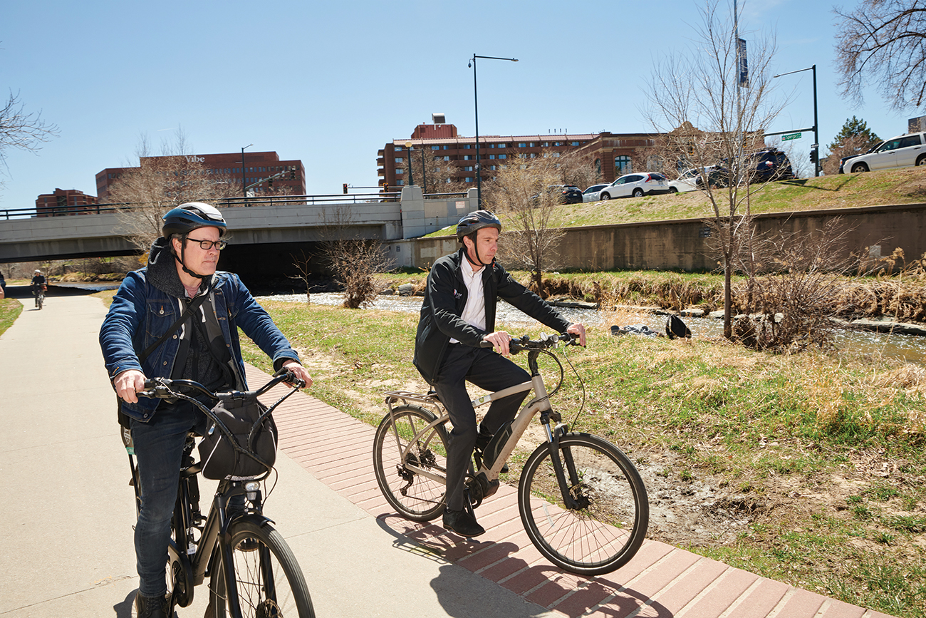 Two men wearing bike helmets ride electric bikes on a paved path.