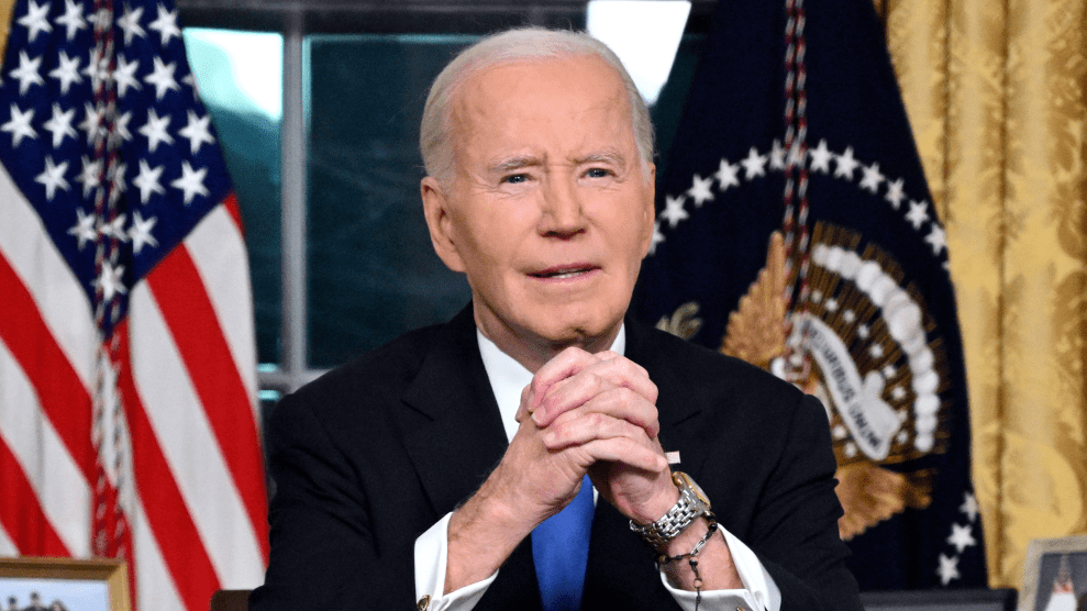 President Joe Biden sits in the Oval Office for his farewell address. Hands clasped. He is wearing a silver and gold watch. The American flag is behind him.