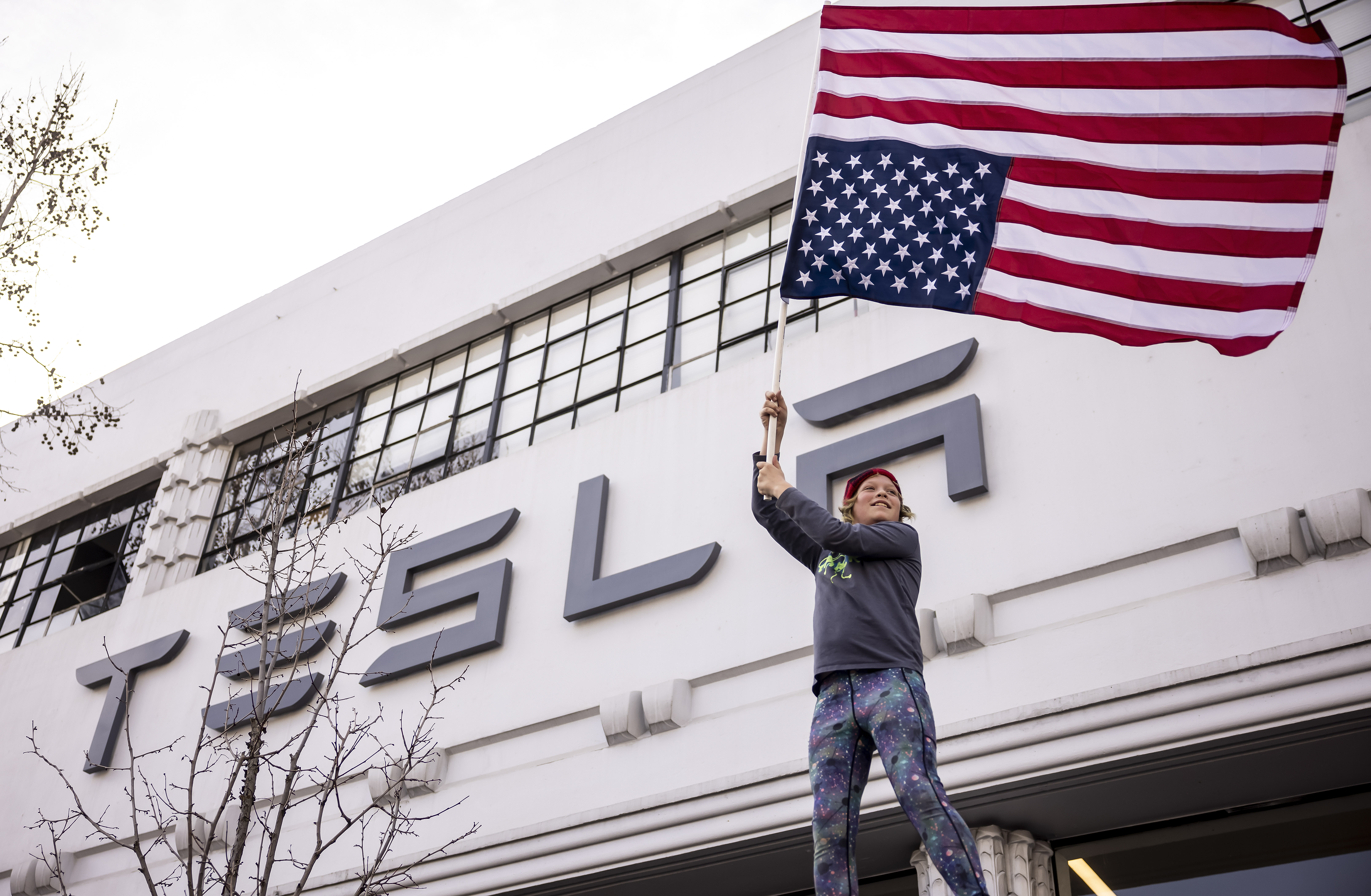 Young person standing below a Tesla dealership sign waving an upside down American flag.