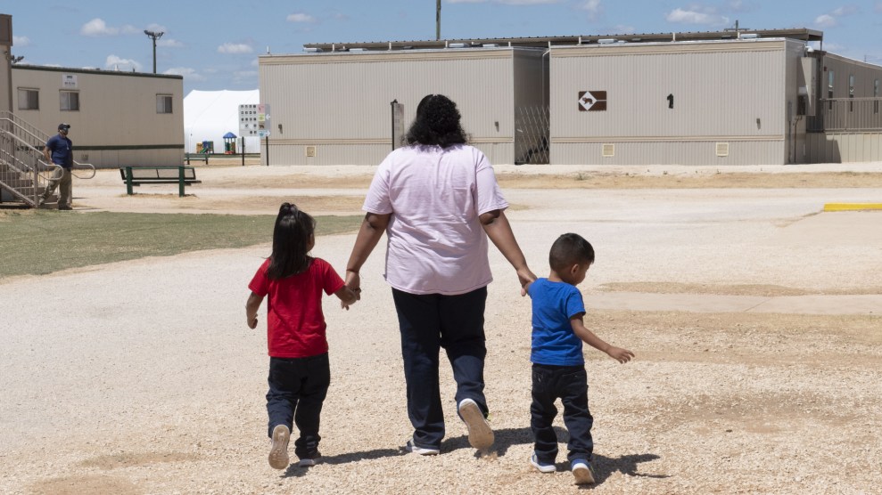 A woman holds hands with two children as they walk across a gravel lot toward industrial-looking one-story buildings.